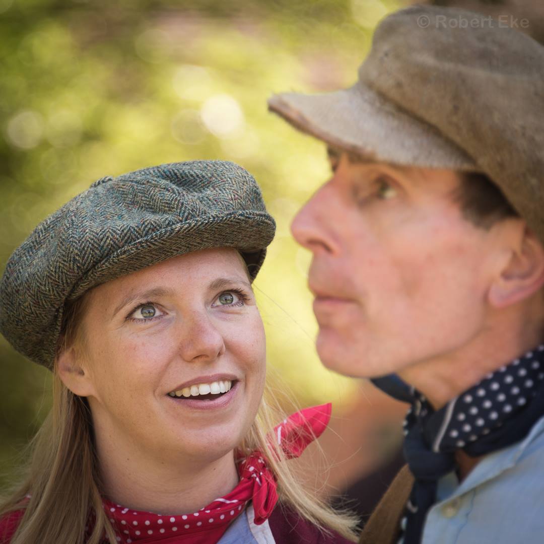 Close up. Left, Gillian Dean as Victoria Stannard wearing flat cap and red neckerchief smiles up a a man, right, wearing a flat cap and blue neckerchief.
