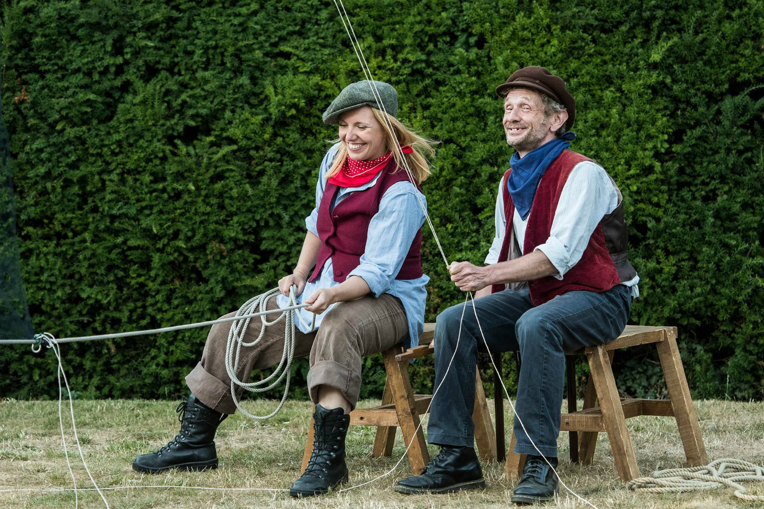 Left Gillian Dean as Victoria Stannard sits on a stool coiling a rope. Right a man sticks on a stoolholding a rope that is coming from above him. Both are smiling.