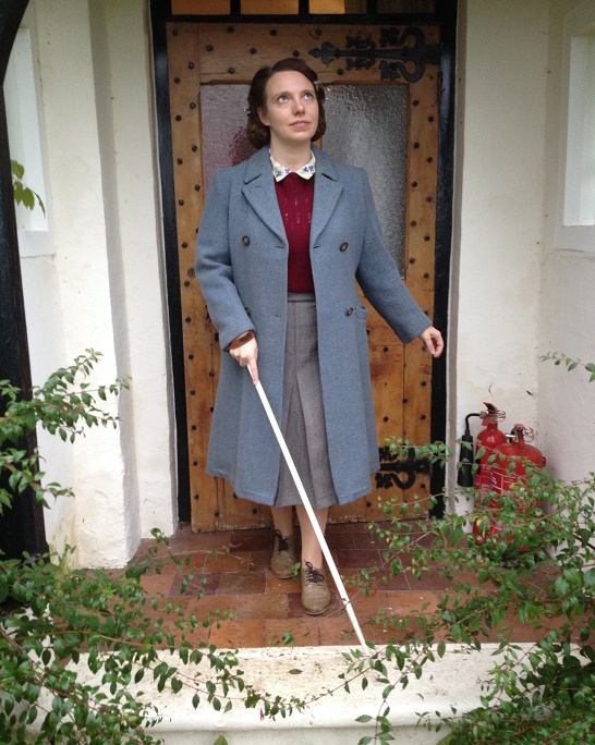 Colour picture of Gillian Dean in front of a cottage door. She is holding a white cane and wearing 1940s clothes