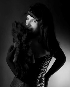Black and white picture. Gillian Dean looking over her shoulder, smiling through a black feather fan. She is wearing a corset.