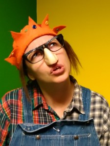 Gillian Dean as Wark/The Ugly Girl. She is looking totally baffled, wearing a Groucho Marx nose and glasses set, an orange feltjesters hat and mismatching checked shirt.