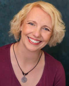 Head and shoulders shot of Gillian. She is a white woman in her early 40s with a tousled blonde bob, and grey green eyes. In this image she is wearing a mauve, v-necked, Henley top, a silver spiral necklace on a black cord, and she is smiling brightly.