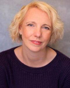 Head and shoulders shot of Gillian. She is a white woman in her early 40s with a tousled blonde bob, and grey green eyes. In this image she is wearing a purple, ribbed-knit jumper, and is looking happy and thoughtful.