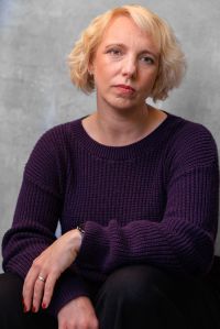 Upper body shot of Gillian. She is a white woman in her early 40s with a tousled blonde bob, and grey green eyes. In this image she is sitting with knees just visible where they are resting on step. Her elbows are resting on her knees, her left wrist is crossed over her right. She is wearing a deep purple, ribbed-knit, crew-neck jumper and black trousers. Her expression is sad and wistful.