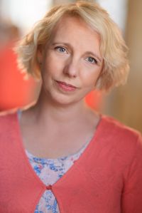 Head and shoulders shot of Gillian. She is a white woman in her early 40s with a tousled blonde bob, and grey green eyes. In this image she is wearing a peach cardigan over a white top printed with small pink and blue flowers. She is looking kind and approachable with a hint of a gentle smile.