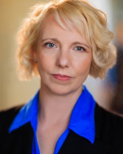 Head and shoulders shot of Gillian. She is a white woman in her early 40s with a tousled blonde bob, and grey green eyes. In this image she is wearing a bright blue shirt under a black waistcoat. She is looking direct and calmly efficient.