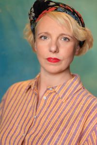 Head and shoulders shot of Gillian. She is a white woman in her early 40s with a tousled blonde bob, and grey green eyes. In this image she has her hair tied in a black, floral headscarf, has red lipstick on, and is wearing a lilac and peach striped shirt. She is looking slightly pensive. The image has a 1940s feel.