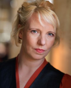 Head and shoulders shot of Gillian. She is a white woman in her early 40s with a tousled blonde bob, and grey green eyes. In this image she has her hair half pinned back, and is wearing an orange satin shirt under a black leather waistcoat. She is looking playful and mysterious.