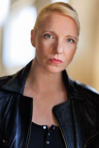 Head and shoulders shot of Gillian. She is a white woman in her early 40s with a tousled blonde bob, and grey green eyes. In this image she is has her hair tied back so only the short, side-sweeping fringe is visible. She is wearing a black Henley top under a battered leather jacket. She is looking direct and severe.